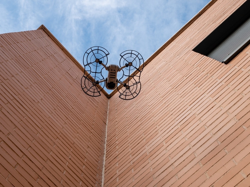 A worm's eye view of a drone with propeller guard flying beside a building